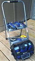 Two-step foldable safety ladder with hand tools arranged on the steps and blue-black Kobalt tool carrying case on the floor next to ladder.