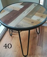 Round auxiliary table with a multi-color wood plank top and black metal legs on hardwood floor.