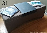 Photo of rectangular beige upholstered ottoman storage bench with tufted cushioned top, curved bottom with nailhead trim, dark wooden legs, dark wood auxiliary tray, and two books on top