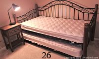 Sofa bed with black metal frame, brown post corners, white mattress on top and white pop-up trundle mattress below, labeled 26. Wooden nightstand with carved drawer front and lamp on top next to sofa.