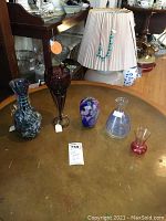 Several small art glass vases arranged on a large brass or bronze round table surface, showing different colors, shapes, and patterns including blue, purple, red, and clear glass.