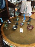 Top-down view of the 11 glass items arranged on a round table including glasses, vases, figurines, and bowl showing color and shape details.