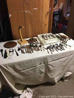 Photo shows an assortment of flatware including numerous forks, spoons, and knives laid out on a table. Some flatware pieces are in a white organizer tray. Background items are not included.