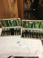 Two open boxes of German silver flatware showing the arrangement of small forks and spoons inside with green satin-lined lids and black fabric bases.