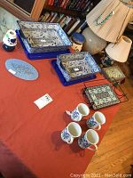 Blue and white Polish pottery casseroles with lids, glass tray in metal carrier, four decorated mugs, and votive holder with shade on a red cloth surface.