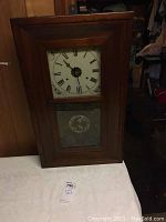 Full view of large wooden wall clock with Roman numeral face and decorative lower glass panel.