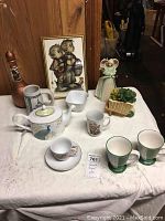Front view of lot items including peacock teapot, mugs, stein, angel figurine, and framed Hummel print.