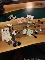 Photo showing multiple pairs of earrings laid out on wooden surface, various styles including studs, hoops, dangles, and textured metal earrings.