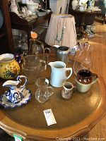 Nine pitchers arranged on a round wooden table including glass, ceramic, and aluminum varieties.