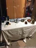 Full view of all lot items arranged on covered table showing plates, glasses, mugs, brass pitchers, redwood burl piece, clothes brush, and other items.