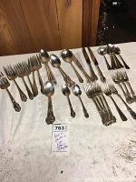 Mixed silver plated flatware including forks, knives, tablespoons, teaspoons, and salad forks arranged on a white tablecloth. Various patterns by Reed and Barton and Wm. Rogers.