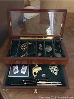 Wooden jewelry box with transparent glass lid open showing multiple bracelets, watches, brooches, cufflinks and pens in lined compartments
