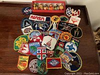Full view of various embroidered patches spread on a wooden surface, showing multiple colors and designs including NASA, National Historic Site, and state emblems.
