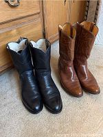 Two pairs of men's Justin cowboy boots, one black and one brown, placed side by side on carpet in front of a wooden dresser.