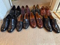 Six pairs of men's dress shoes arranged in two rows on carpet, showcasing styles, colors, and wear.