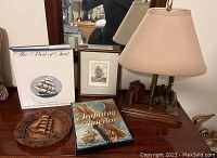 Overview of sailing-related items including framed etching, two books, carved wooden plaque, and nautical lamp on wooden table.