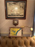 Framed ship print with brass finish adjustable wall lamp, wooden nautical tray with inset map, and small framed nautical engraving all displayed above a leather couch