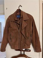 Front view of a brown leather jacket hanging on a wooden hanger showing zipper front, two snap-button pockets, and the folded collar.
