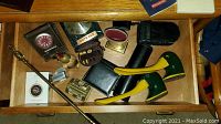 Photo of an open drawer containing various vintage items including two pineapple-shaped shoe horns, duck-shaped shoe horn/lint brush, two clocks, leather wallet, brass vessel, and other small objects.