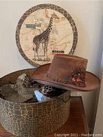 Vintage brown leather hat with wide brim and decorative braided band with feathers and branded pins, displayed on giraffe-patterned box.