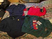 Four vintage men's sweaters laid out on a bed: navy blue crewneck, red and black color-blocked, dark navy polo with collar, and green holiday-themed sweater