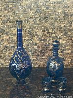 Photo showing two antique blue glass decanters with silver floral detailing and two small matching glasses, displayed against a brick background.