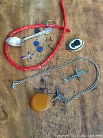 Photo showing various antique jewelry items on wood surface including coral bead necklace, silver spoon, amber pendant necklace, silver crucifix, cameo brooch, blue stone earrings, small blue cross pendant, and silver ring.