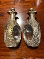 Pair of antique silver metal oriental dragon decanters placed side by side on wood surface showing dragon motifs and clear glass insets.