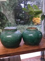 Pair of green glazed antique Chinese jars on wooden surface with glass window and garden background, one lid missing.