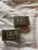 Two rectangular silver match holders laying on fabric with visible detailed coat of arms on front faces.