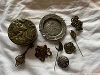 Photo showing grouping of 8 small Art Nouveau items including a round trinket box, a small dish, several decorative pins and brooches, displayed on white fabric.