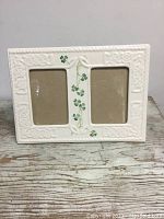 Front view of white porcelain double picture frame with two vertical openings and Celtic embossed detailing along edges, with green shamrock hand-painted accents in the center divider.