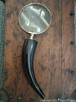Magnifying glass with a curved dark horn handle placed on a wooden surface, showing the round glass lens with metal frame.
