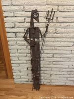 Front view of large iron yard ornament humanoid figure with trident against white brick wall, showing full height and detail of iron construction.