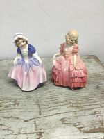 Front view of two Royal Doulton porcelain figurines, one in purple and pink dress with bonnet, the other in pink layered dress