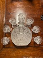 Top-down view of crystal decanter with stopper surrounded by six matching crystal glasses arranged in a circle on a wooden surface.