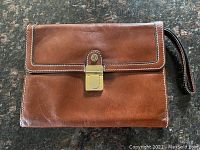 Closed leather wallet showing front brown leather with gold-tone clasp and attached wrist strap.