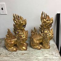 Pair of gold-painted Thai temple dragon statues with colorful mirrored glass decorations, shown side by side on a wooden surface against a white wall.