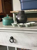 Photo showing black ceramic vase, teal lidded bowl, and four small Chinese miniature porcelain cups with bird and floral decoration and two plain cups placed on a white table.