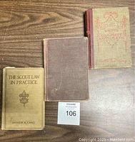 Three antique hardcover books displayed vertically on wood table, showing front covers; titles visible on two books.
