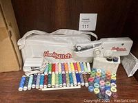 Photo showing white zip-up tote bag with Handy Stitch logo, handheld sewing machine, power adapter, various thread spools in many colors displayed in front on a surface.