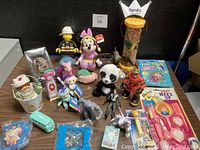 Overview photo of assorted vintage and new toys, figures, plush, and playsets on table surface against black background with lot number card