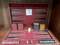 Opened backgammon board showing red felt playing surface with black and brown points, multiple playing pieces, dice, and four cup holders.