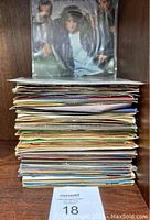 Stack of a large number of sleeved 45 RPM vinyl records on a wooden shelf.