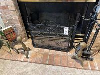 Photo showing black metal fireplace screen with mesh and decorative scrollwork in front of brick fireplace, with metal andirons positioned on brick hearth.