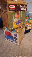 Two images showing boxes of Little Tikes Toddler Swing and Deluxe Booster Seat stacked on carpeted floor near colorful cushioned item.