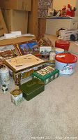Photo showing an assortment of decorative tins arranged on a carpeted floor near cardboard boxes and other packing materials, including holiday-themed and vintage-style tins.