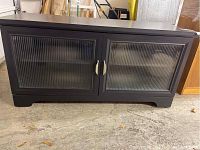 Front view of the black wooden low cabinet showing vertically ridged glass doors and silver handles, on a concrete floor.