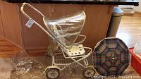 Front and side view of vintage white metal Teutonia stroller with clear attached rain hood and plaid umbrella placed beside it