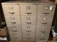 Front view of three 4-drawer vertical file cabinets aligned side by side, light gray color with label holders and recessed handles. Visible wear and scuff marks on surfaces.